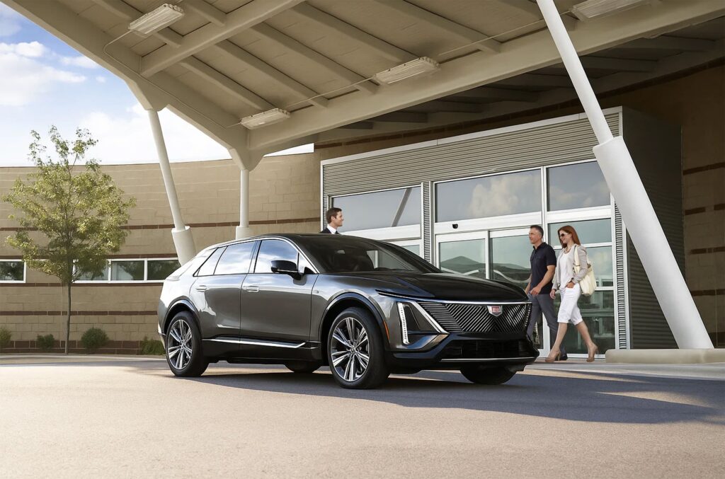 A sleek black SUV is parked under a large canopy outside a modern building, with three professionally dressed people walking nearby.